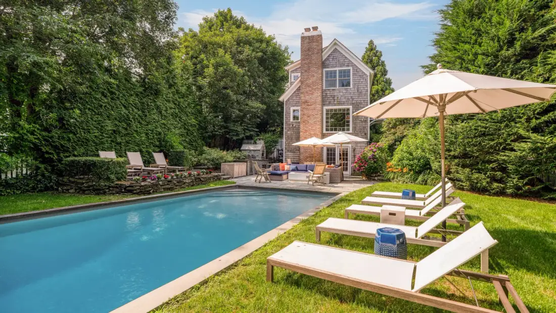 A backyard swimming pool beside a house, with lounge chairs and a large umbrella.