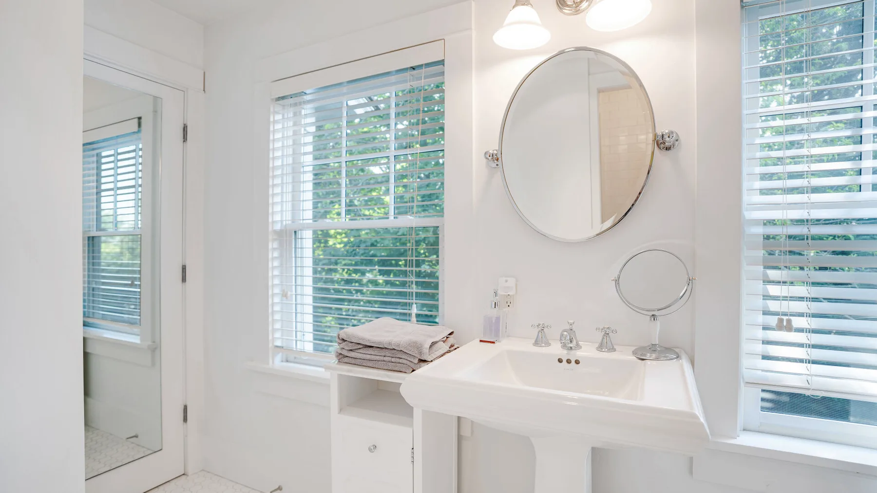 A bright bathroom with a sink, round mirror, and windows with blinds.