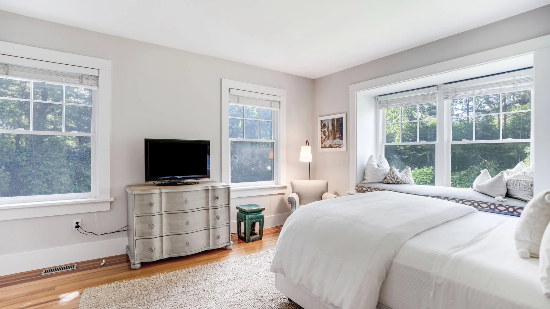 A bright bedroom with a white bed and a wide window overlooking trees.