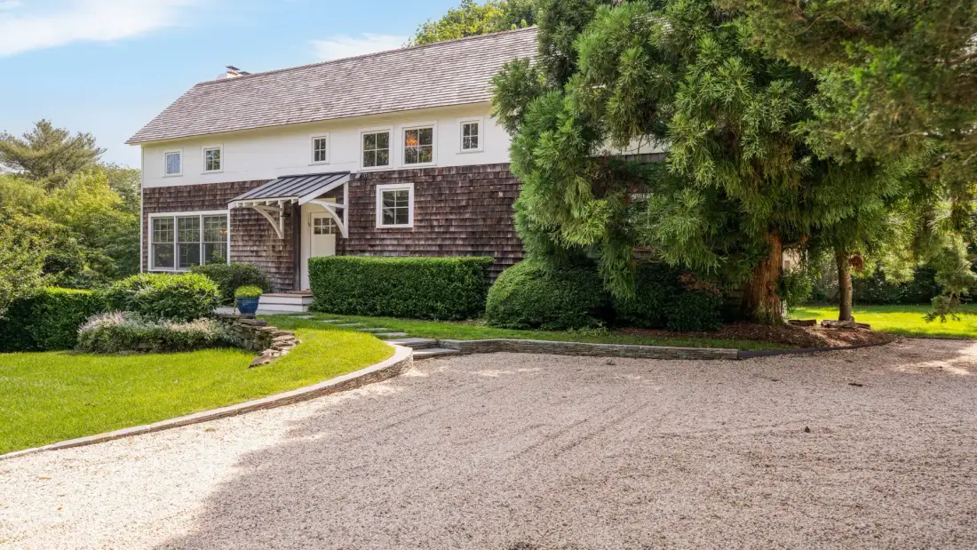 A house with a dark lower facade stands beside a broad gravel driveway.
