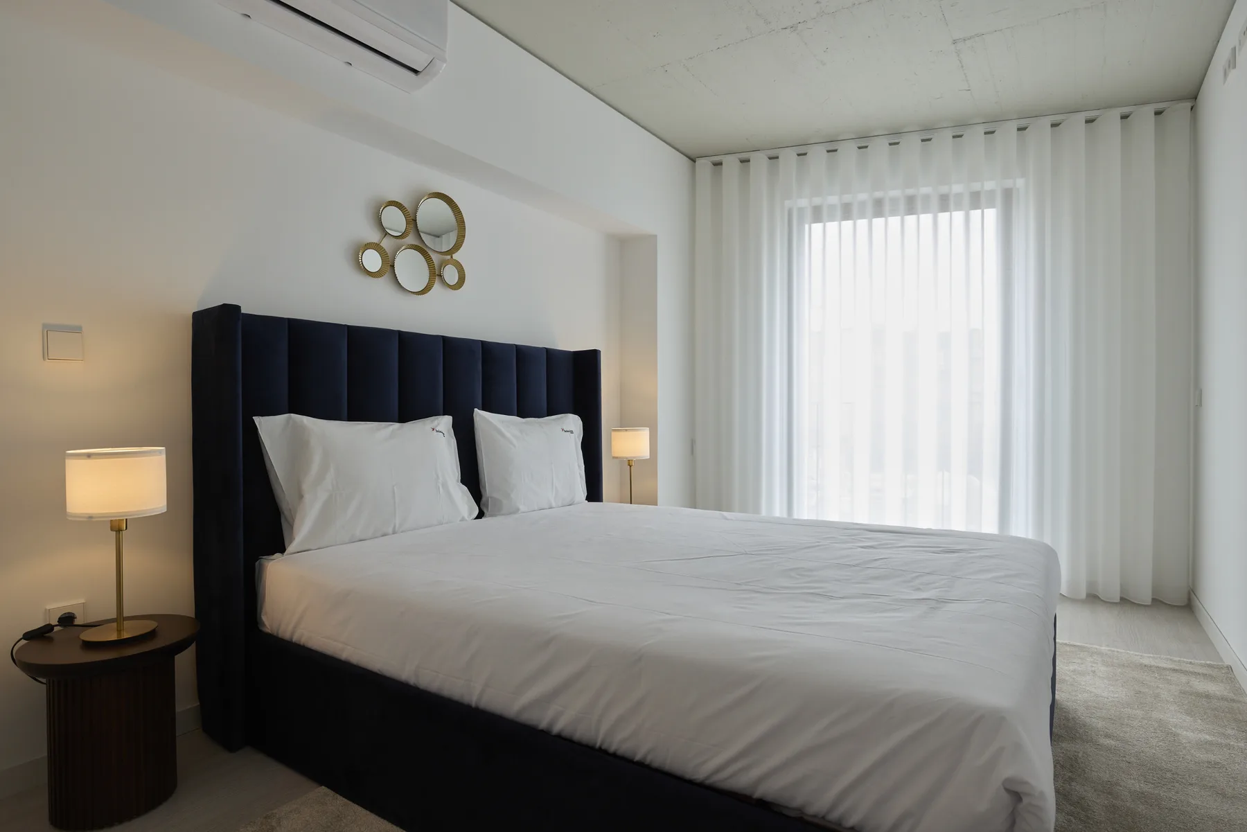 A bedroom with a neatly made bed beside a large curtained window.