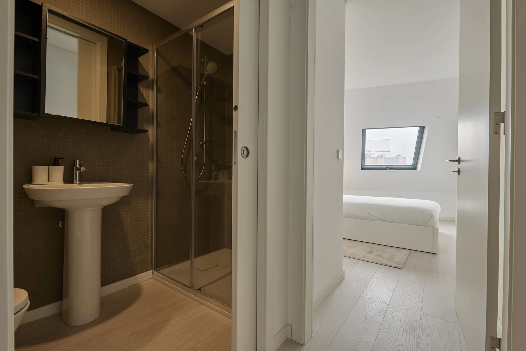 A bathroom with a pedestal sink and glass shower opens onto a bedroom.
