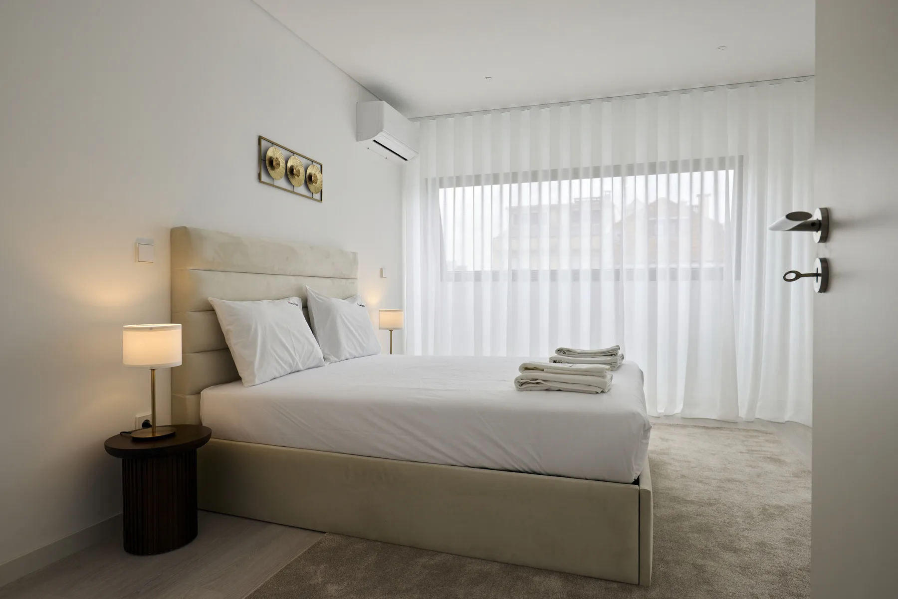 A bright bedroom with a neatly made bed beside a large curtained window.