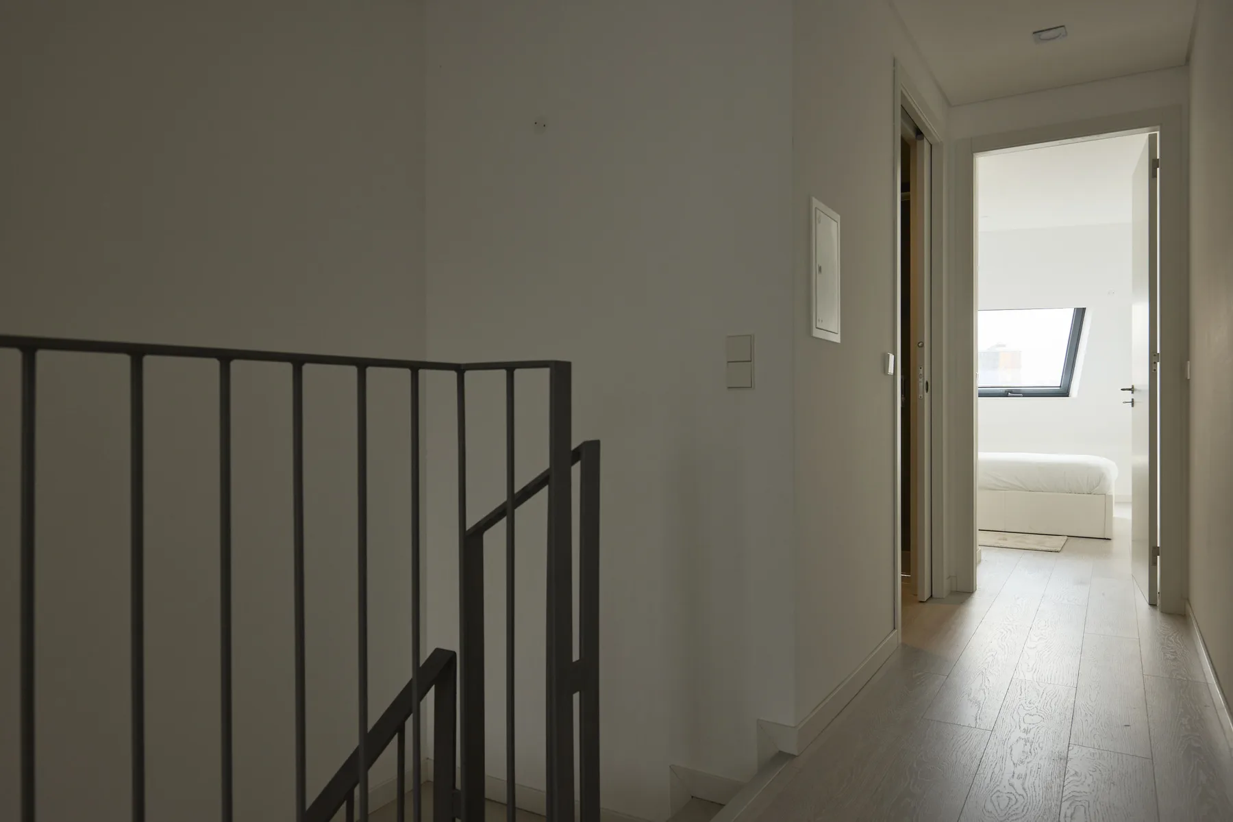 A hallway leads toward a bright bedroom, with a dark railing in the foreground.