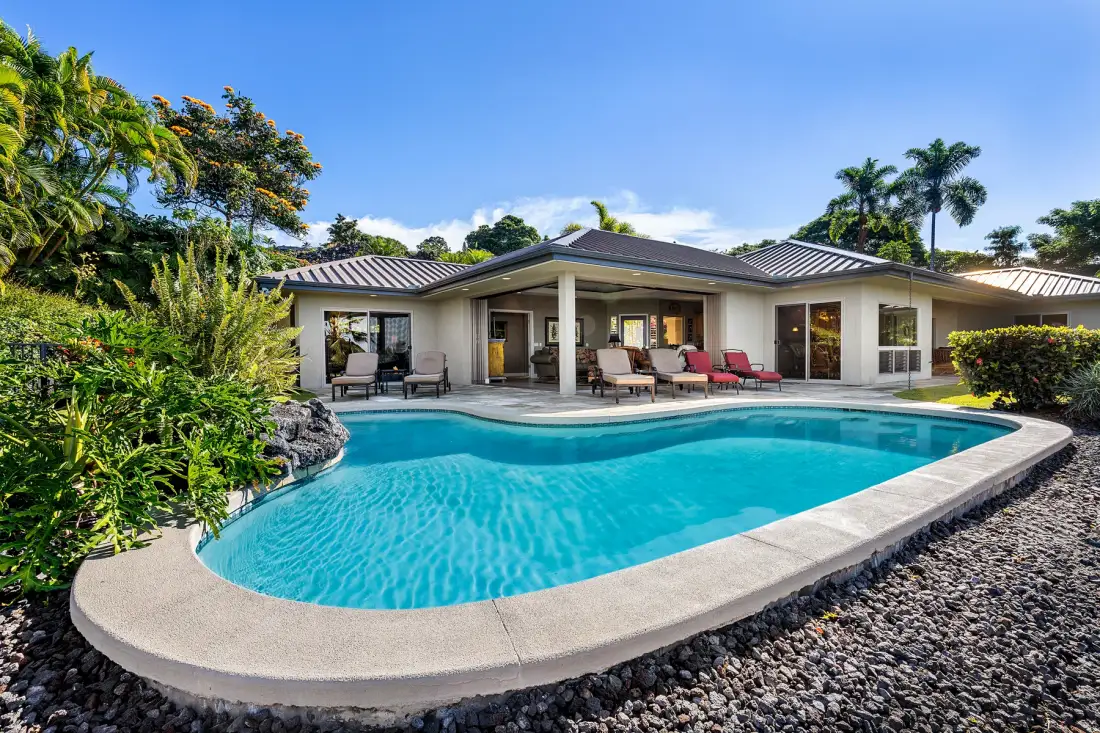 A swimming pool curves in front of a single-story house surrounded by tropical landscaping.
