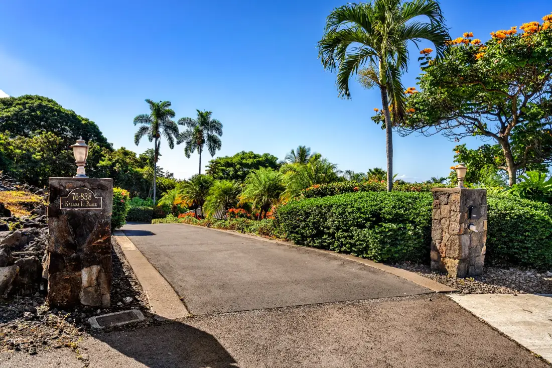 A paved driveway entrance runs between stone pillars, with tropical plants and trees.