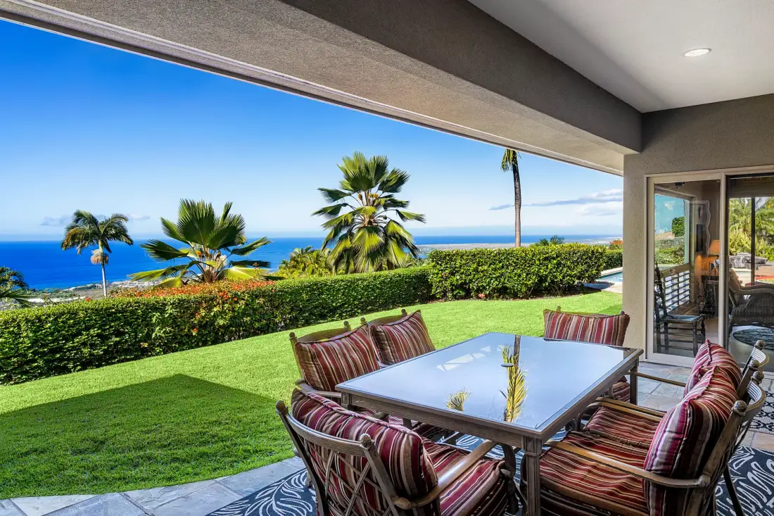A covered patio with cushioned seating and a table overlooks a green lawn and ocean.