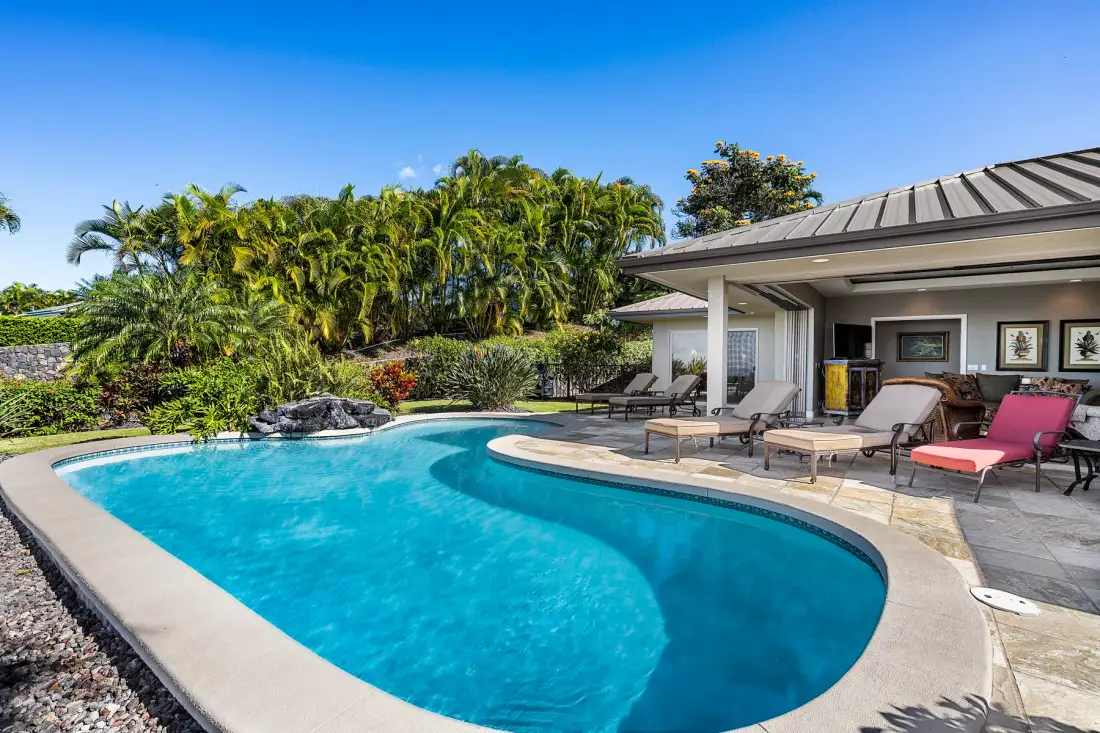 A curved swimming pool sits beside a patio with outdoor seating and tropical landscaping.