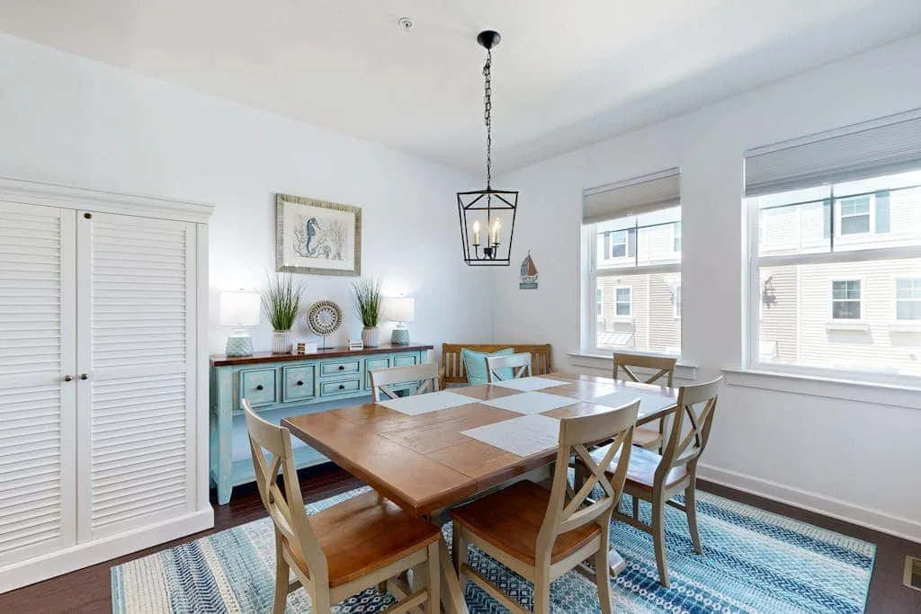 A bright dining room has a wooden table, matching seating, and large windows.