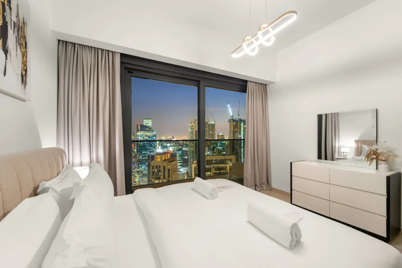 Un dormitorio luminoso con una cama junto a grandes ventanales y luces de la ciudad.