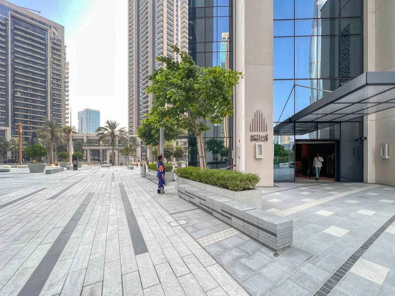La entrada de un edificio moderno da a una amplia plaza pavimentada con árboles y jardineras.