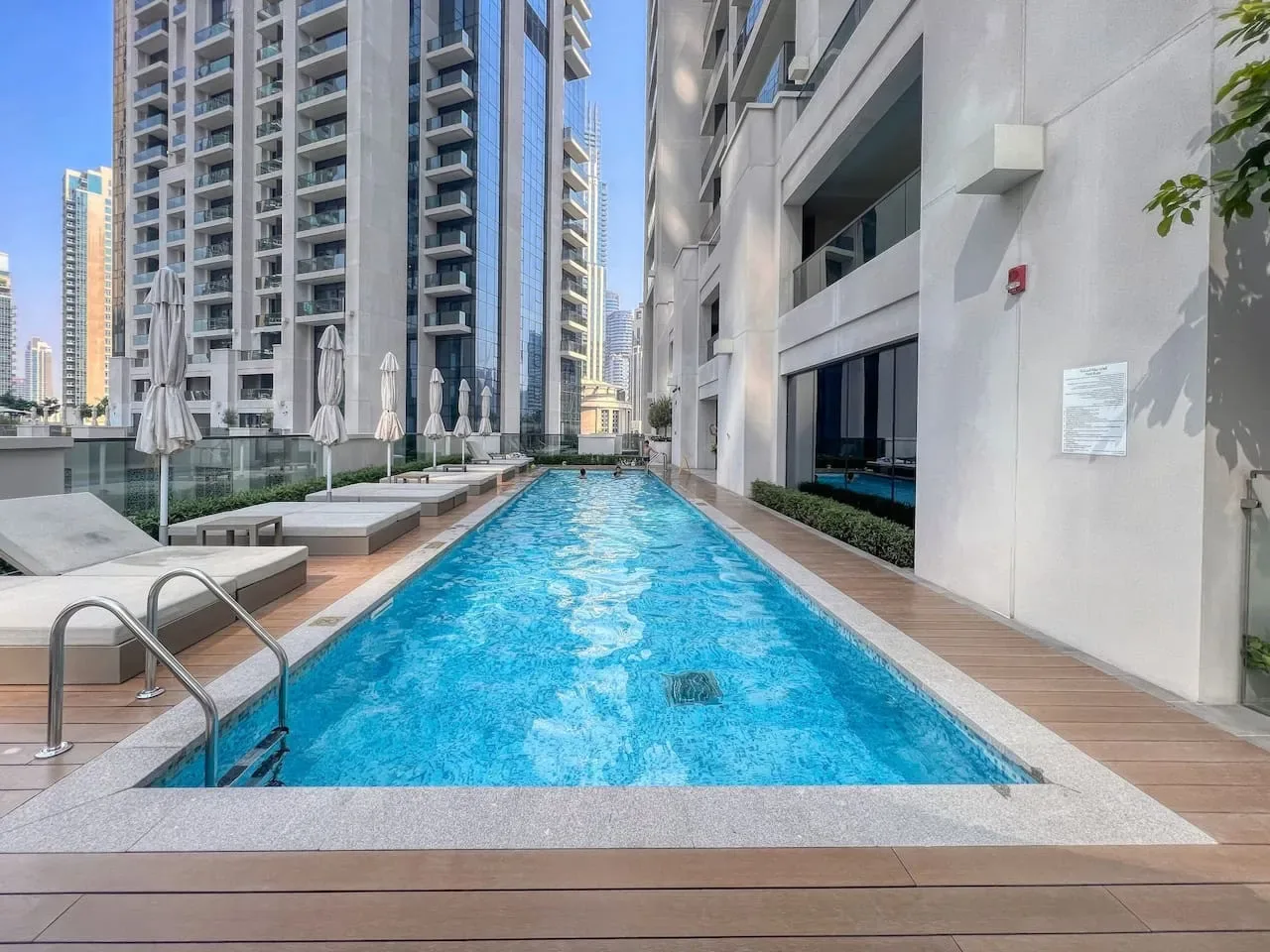 A swimming pool sits between modern high-rise buildings, with lounge seating along the deck.