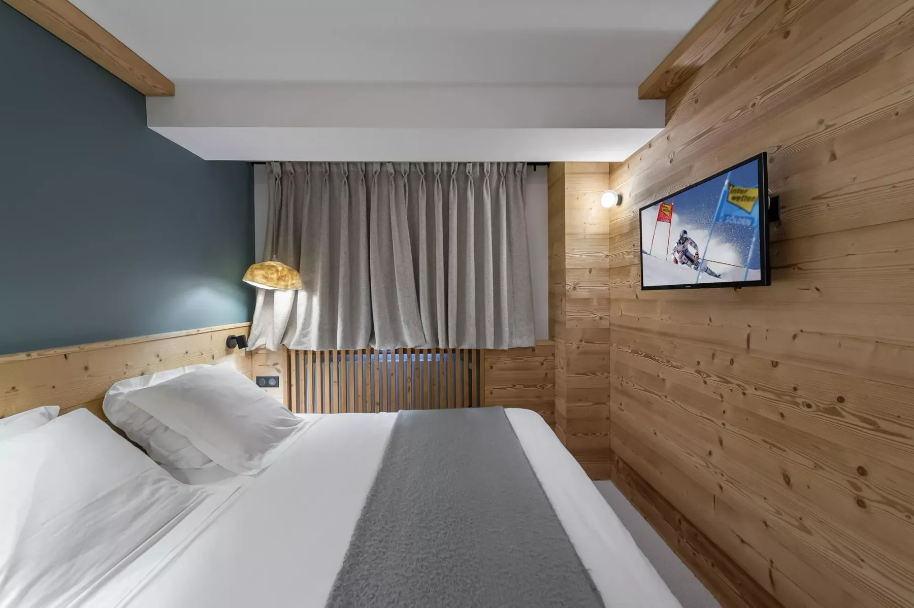 A bedroom with a neatly made bed and a television mounted on the wood-paneled wall.
