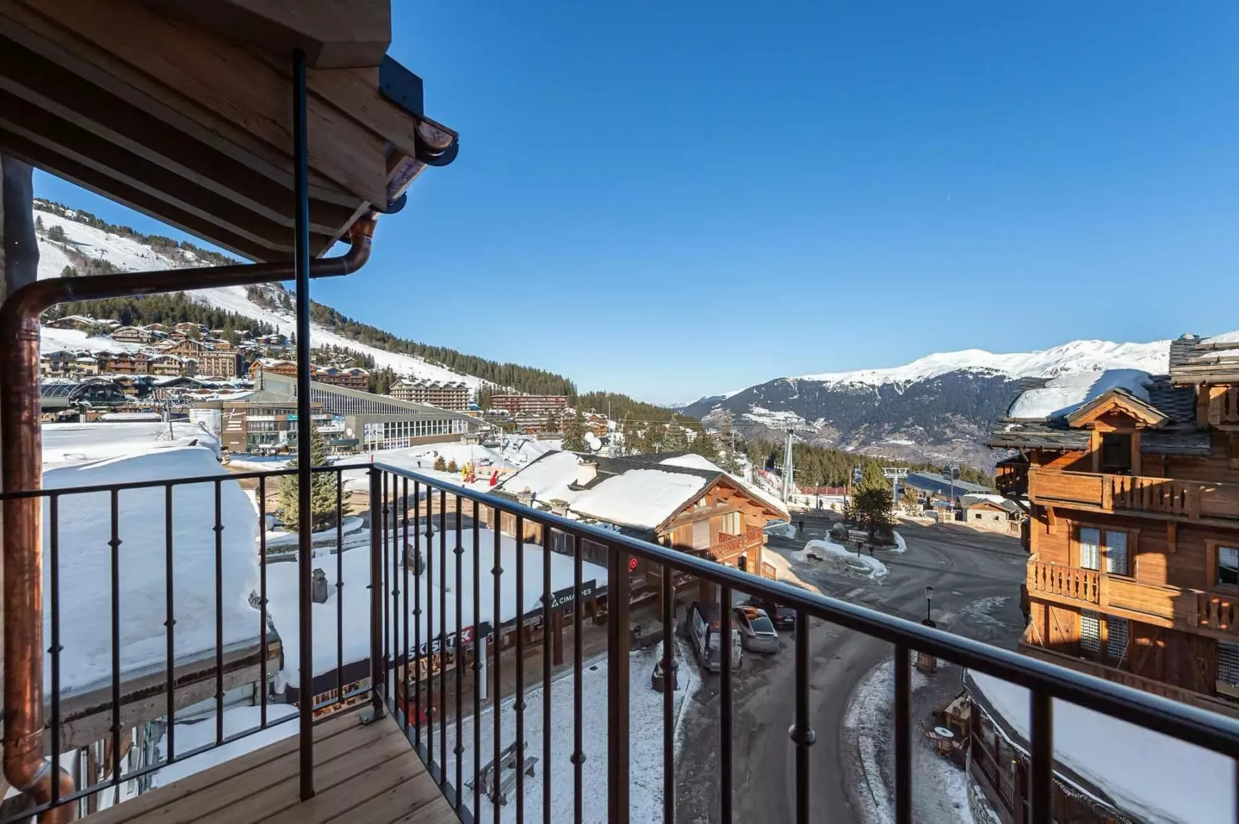 A balcony overlooks snow-covered buildings and a mountain range beneath a clear blue sky.