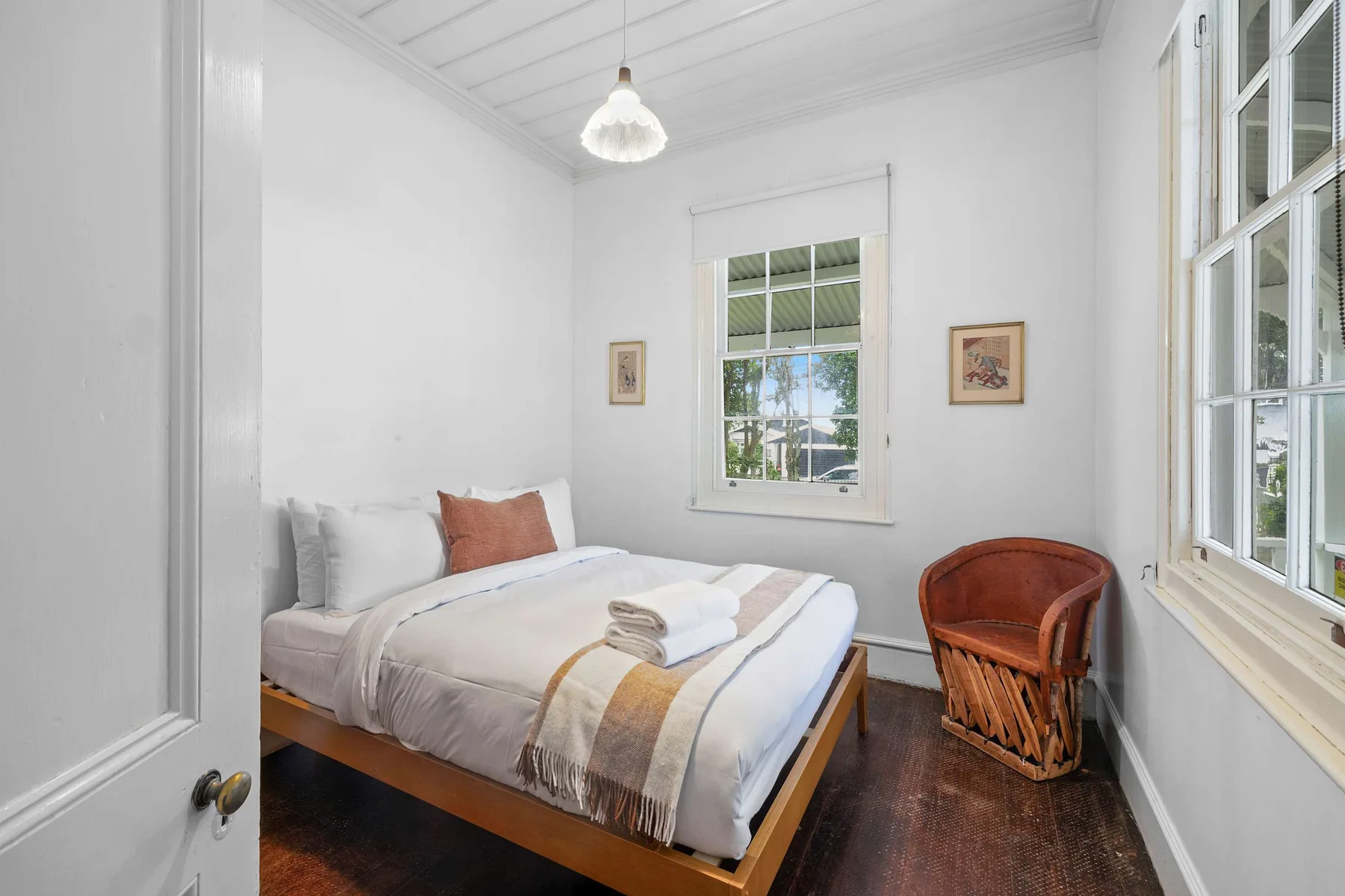 Un dormitorio luminoso con una cama tendida y una silla tejida junto a las ventanas.