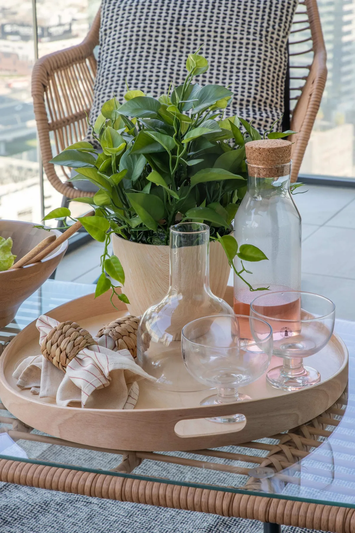 A glass table holds a round tray with a leafy potted plant and clear glassware.