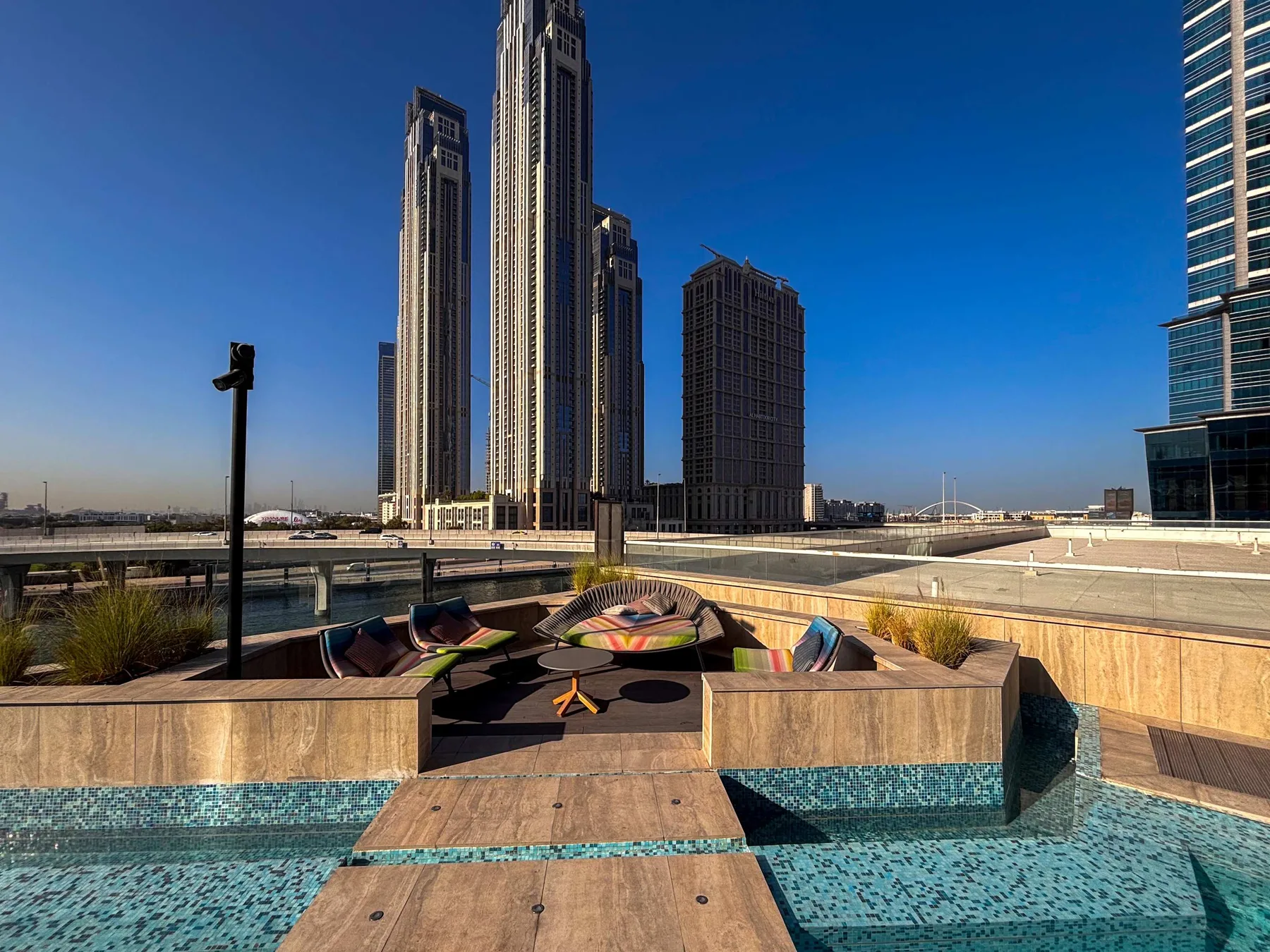 A rooftop pool deck with a wooden lounge platform and tall city buildings in the background.
