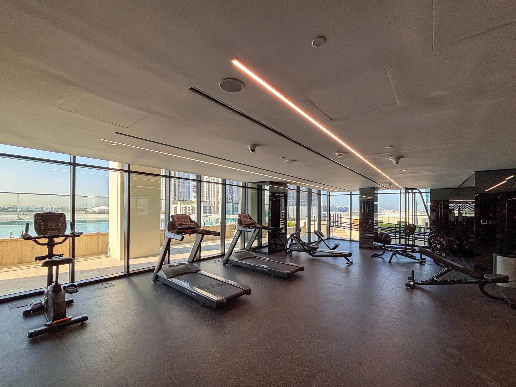 A gym with treadmills and exercise equipment beside large windows overlooking the water.