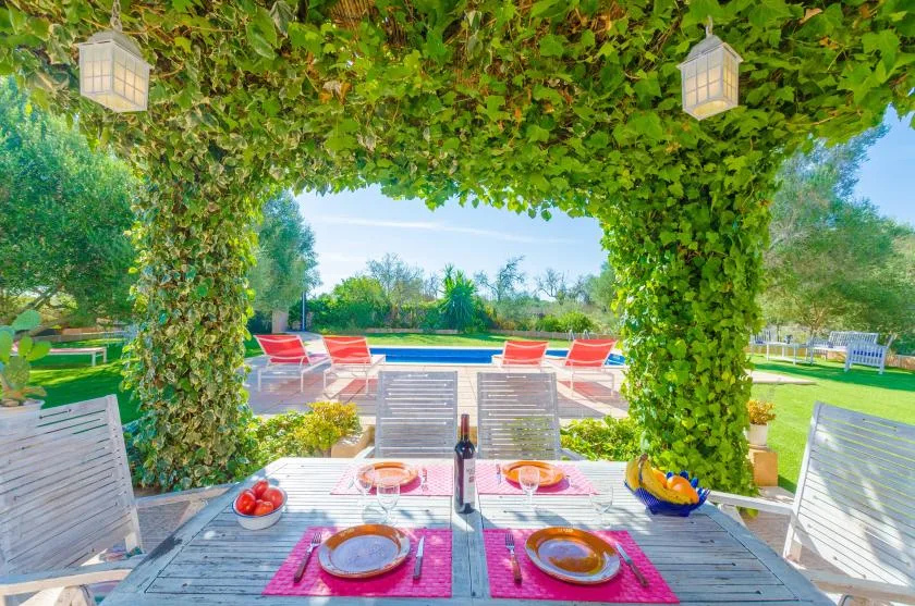 Una zona de comedor al aire libre cubierta de hojas da a una piscina y un jardín.