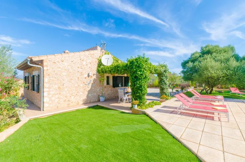Un patio soleado junto a una casa de piedra, con césped verde y asientos reclinables.