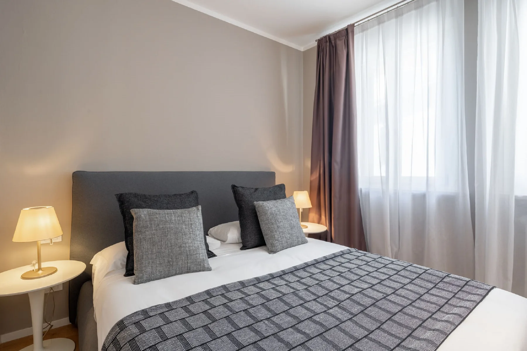 A bedroom with a neatly made bed and sheer curtains over a window.