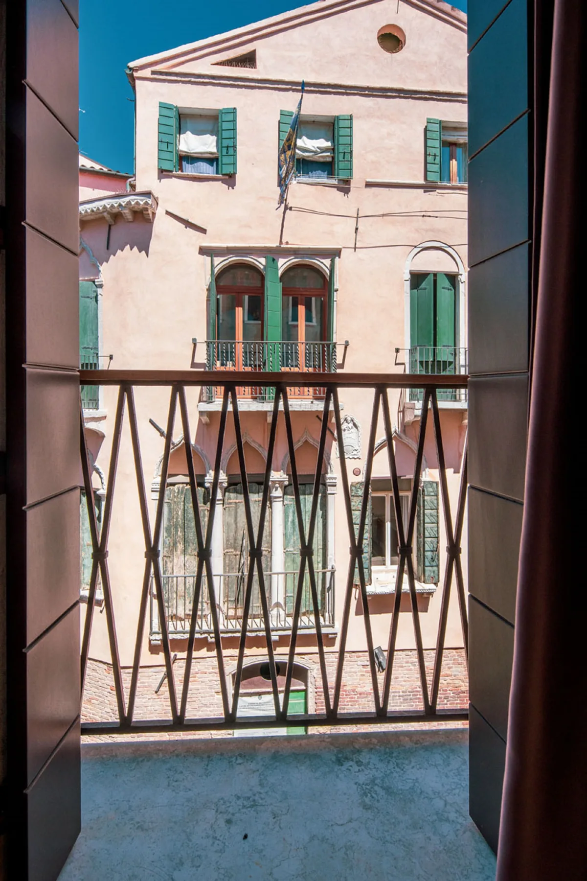 A view of a pale pink building with green shutters across a balcony railing.
