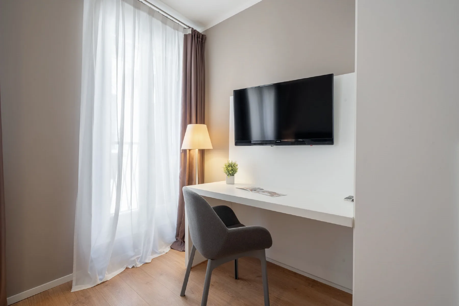A bright bedroom workspace has a desk and wall-mounted television beside a curtained window.
