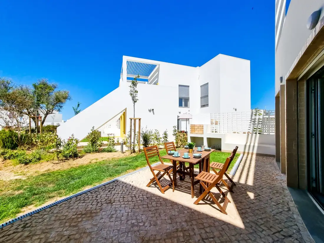 Un patio pavimentado con asientos al aire libre da a un jardín con césped junto a un edificio blanco.