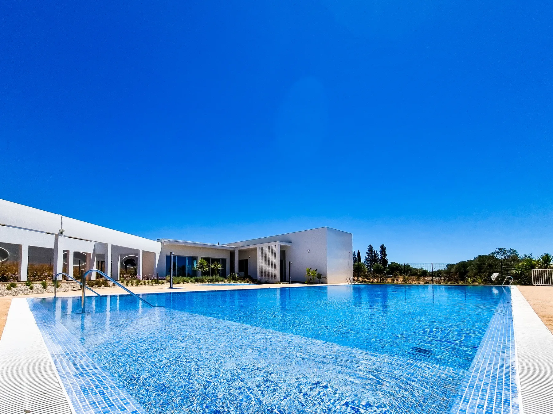 Una piscina está frente a un edificio moderno de poca altura bajo un cielo azul despejado.