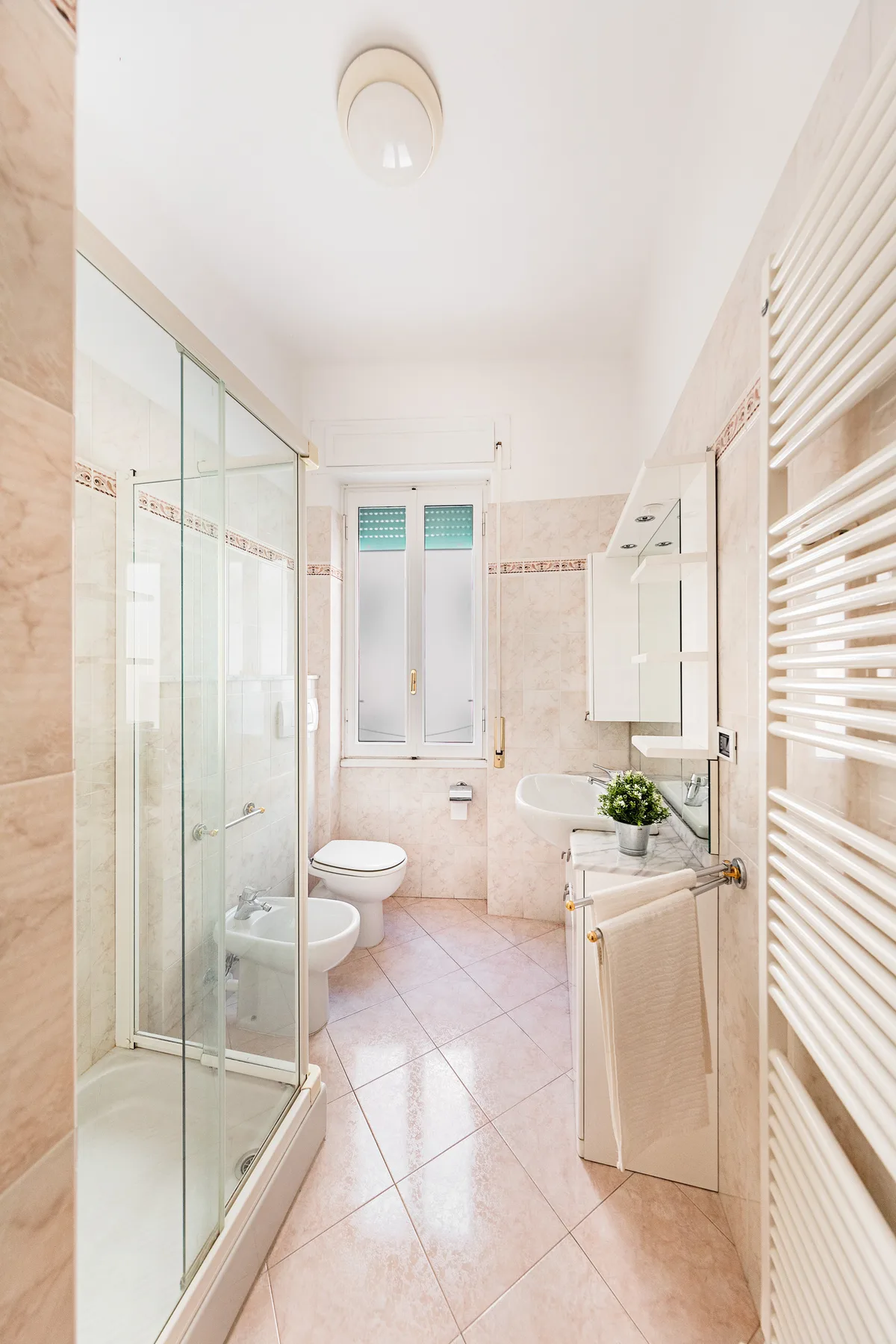 A bright tiled bathroom with a glass shower and a window at the far end.