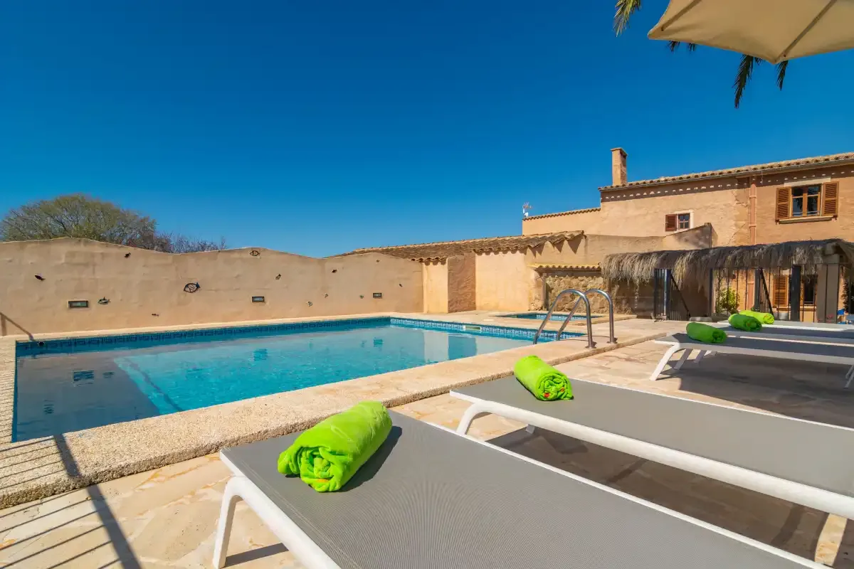 A sunny poolside patio with loungers beside a swimming pool and stone building.