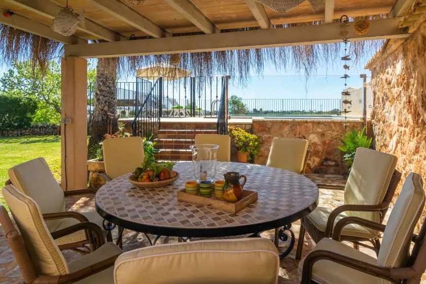 A shaded outdoor dining area features a round table and cushioned seating.
