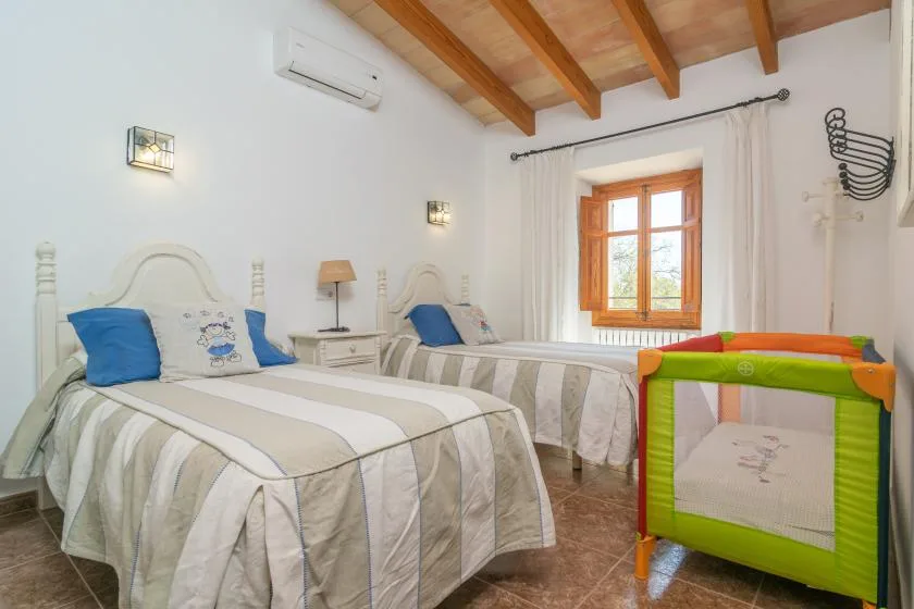 A bright bedroom with two beds and a colorful playpen beside the window.