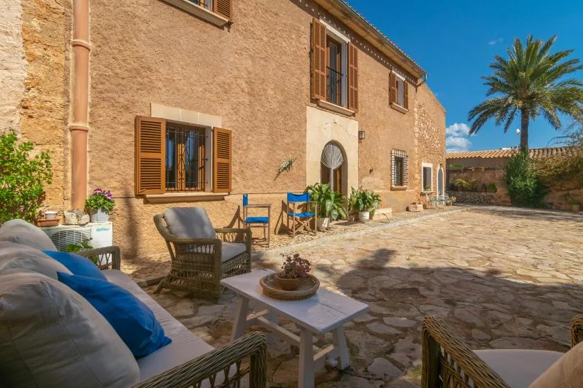 A stone courtyard beside a rustic building, with outdoor seating and a palm tree.