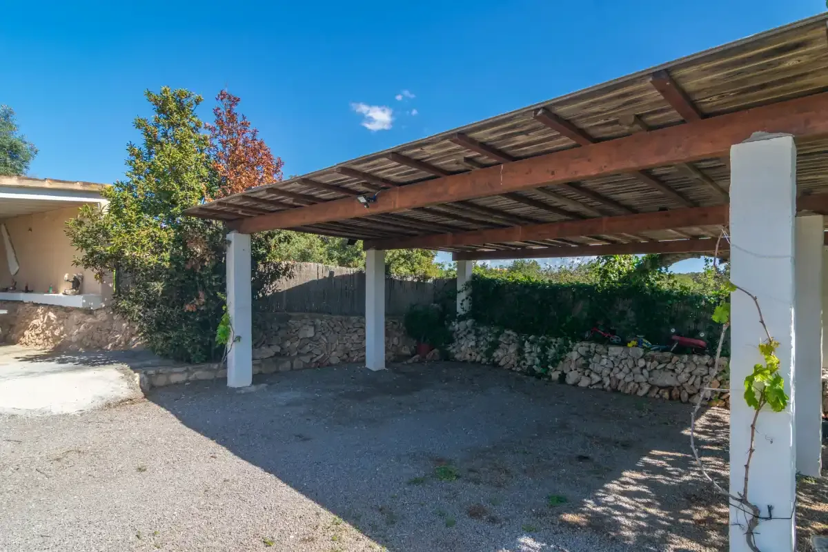 A shaded outdoor parking area sits beneath a wooden pergola beside a stone wall.