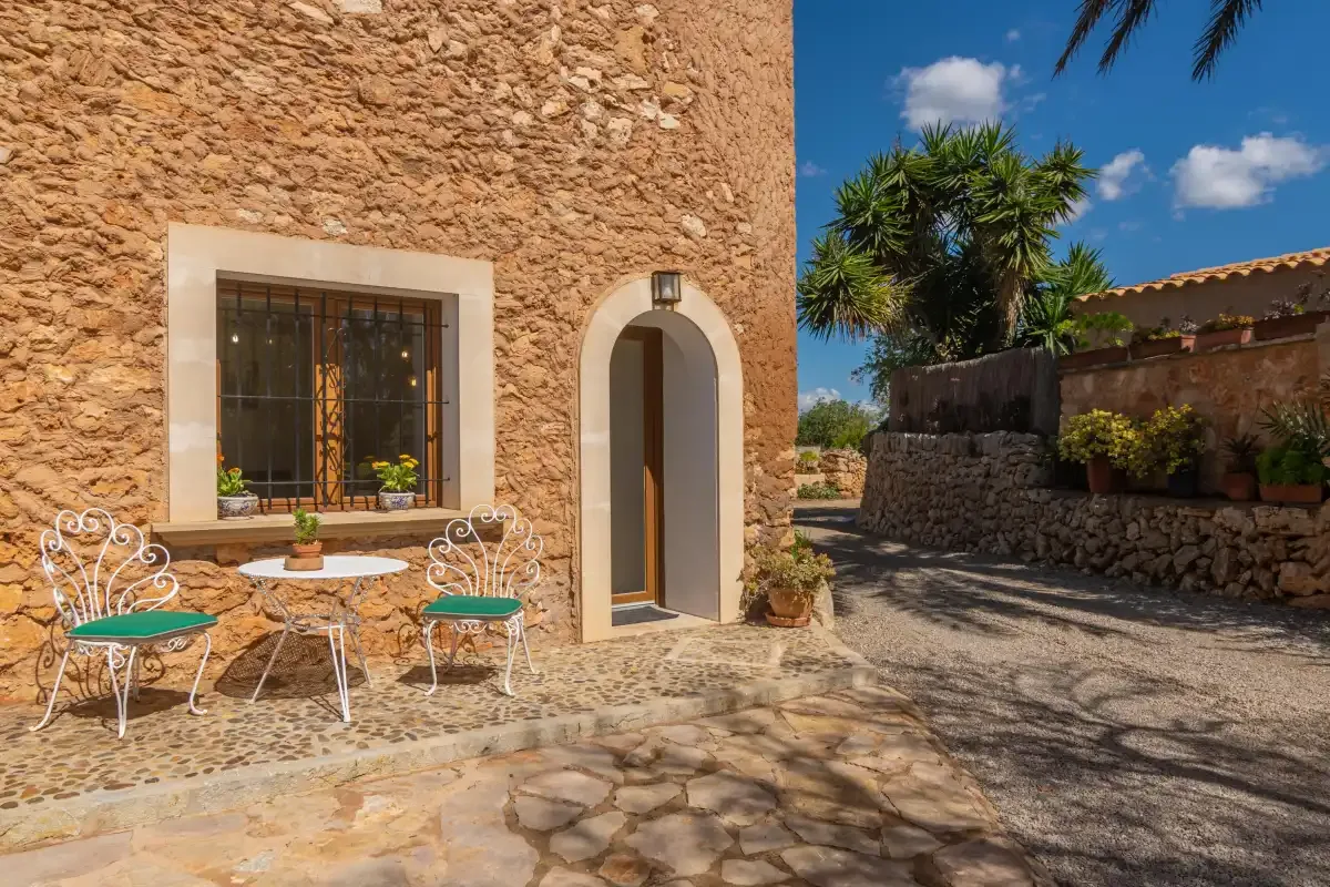 A stone building entrance opens onto a sunny courtyard with a small outdoor seating area.