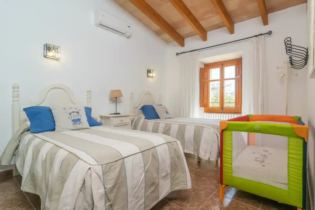 A bedroom with two beds, a window, and a colorful playpen.