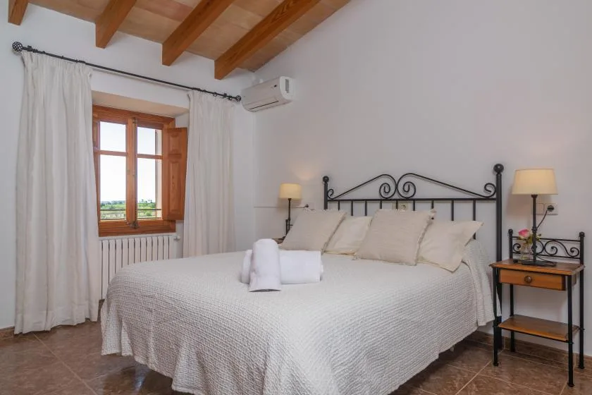 A bedroom with a large bed, a curtained window, and exposed wooden ceiling beams.