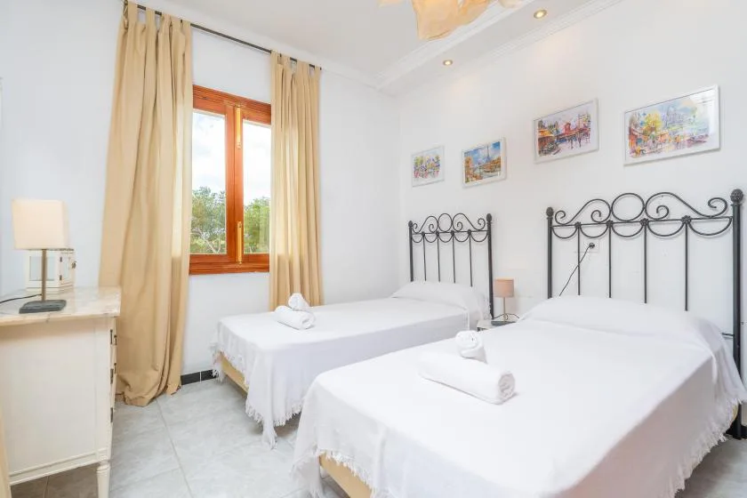 Un dormitorio luminoso con dos camas, cortinas beige y cuadros enmarcados.