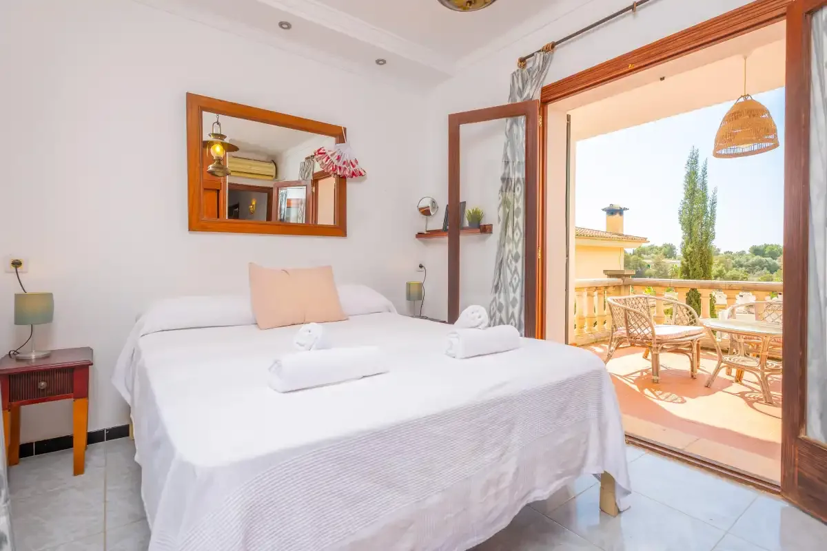 Un dormitorio luminoso con una cama tendida junto a puertas abiertas a un balcón.
