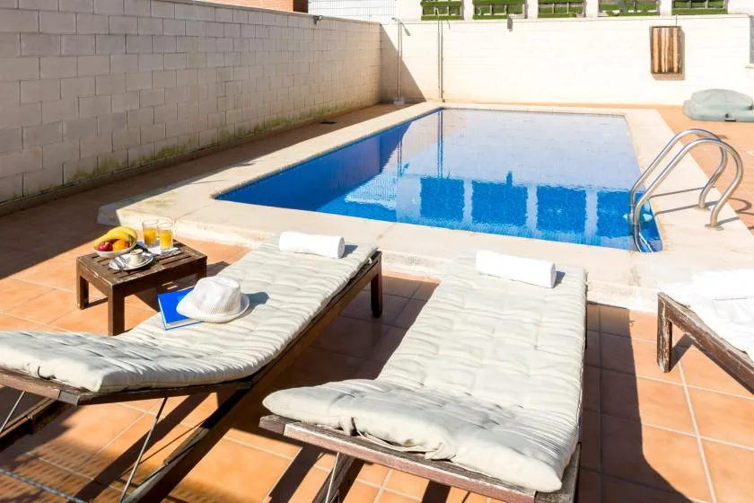 A sunny poolside terrace with lounge seating beside a blue swimming pool.