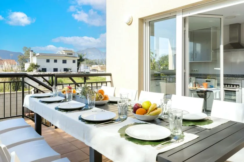 An outdoor dining table is set beside large glass doors, with a sunny view beyond.