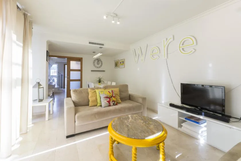 A bright living room with beige seating, a television, and a round yellow table.