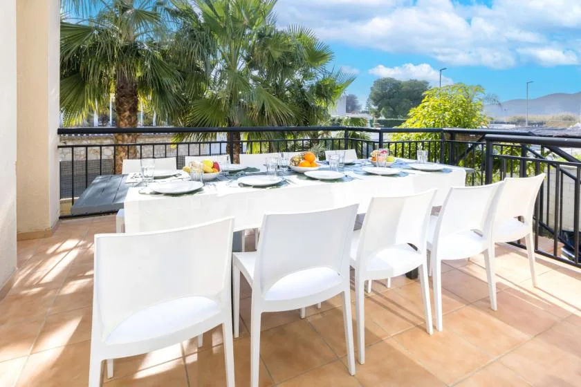 A dining table and seating sit on a sunlit terrace overlooking palm trees.