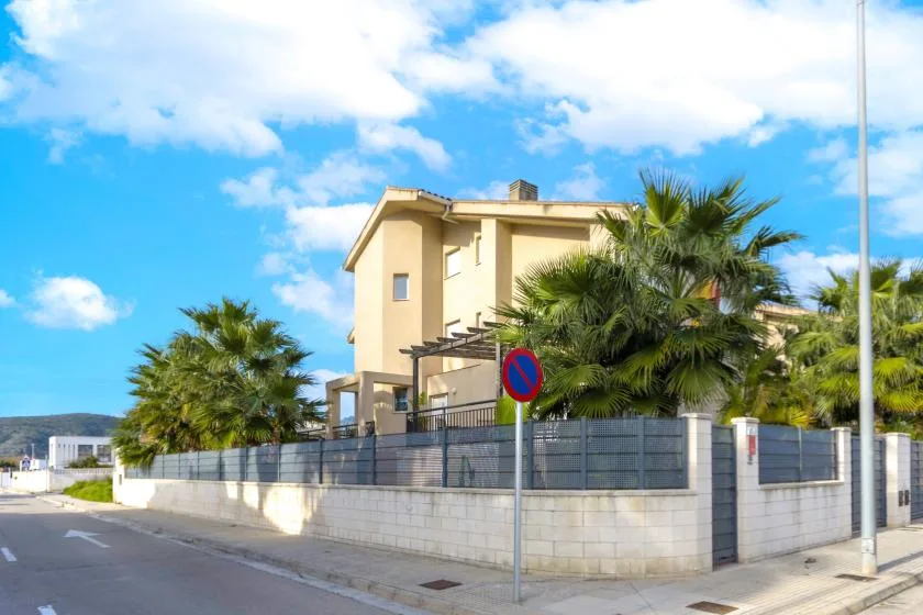 A beige house stands behind a low wall and palm trees beside a street.