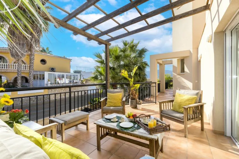 A sunlit outdoor terrace with seating, potted plants, and a pergola overhead.