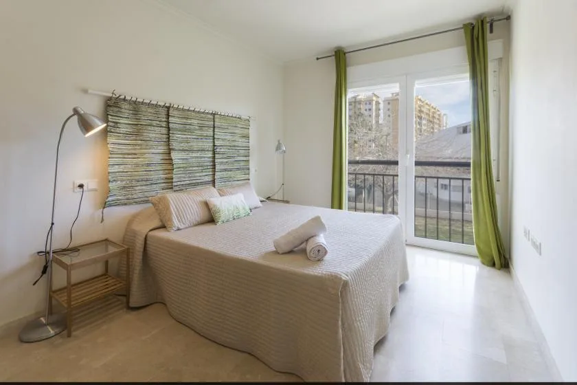 A bright bedroom with a neatly made bed beside glass doors opening onto a balcony.
