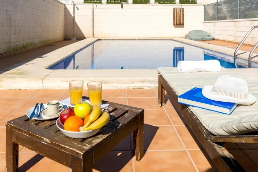 An outdoor poolside patio with a table of fruit and drinks beside lounge seating.