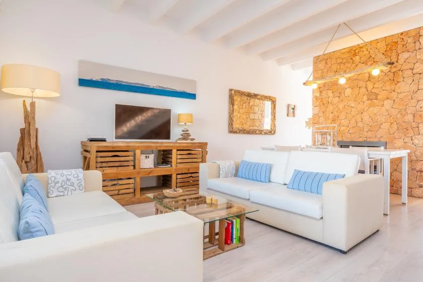 Una sala luminosa con asientos blancos, un mueble de madera para televisión y una mesa de centro de vidrio.