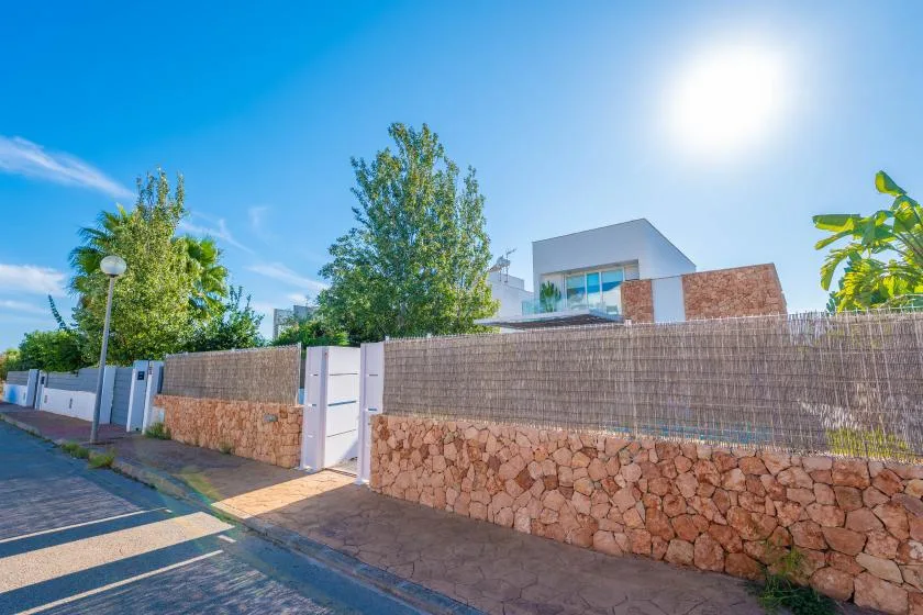 Una casa moderna se alza tras un muro de piedra y malla junto a una calle pavimentada.